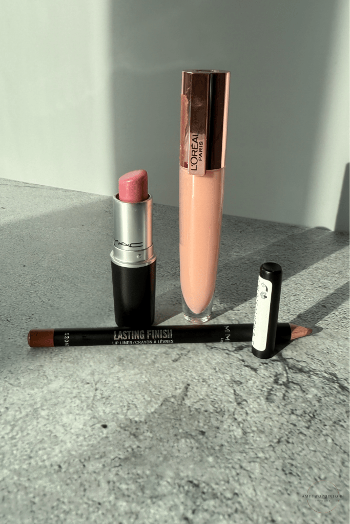 A flat‑lay of a pink‑toned nude lip combo, including a MAC pink‑nude lipstick, a brown Rimmel Cappuccino lip liner, and the L’Oréal Lifter Gloss in the shade Soar. The products are arranged on a neutral surface in soft natural light, showing the tones clearly — a cool‑leaning pink lipstick, a warm brown liner for definition, and a milky pink gloss that gives a smooth, blurred finish