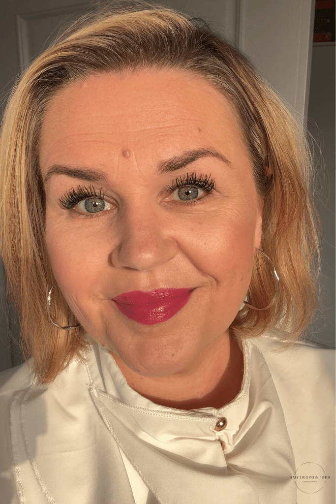 blonde hair and green eyes over 40 showing a lipstick tube in shade 108 fit check Maybelline 8HR Serum Satin Lipstick – Fit Check and the colour on the lips in natural sunlight